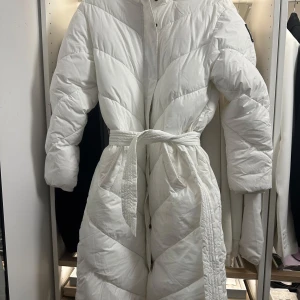 Vit lång pufferjacka från Zink - Säljer en lång, vit pufferjacka från Zink med bälte i midjan och snygg quiltad design. Jackan har långa ärmar, knappar framtill och en cool patch på ärmen. Perfekt för kalla dagar när du vill vara både varm och stilren.
