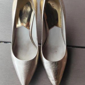 Guldiga pumps från Michael Kors - Snygga pumps i metallic guld från Michael Kors med spetsig tå och hög klack. Skorna har en elegant struktur i materialet och insida i guldton. Perfekta för dig som vill sticka ut med stilrena detaljer och lyxig känsla.