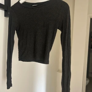 Mörkgrå ribbad långärmad tröja - En enkel och stilren mörkgrå långärmad tröja med ribbad struktur. Tröjan har en rund halsringning och är något croppad i modellen, vilket ger en modern och avslappnad look. Perfekt att matcha med jeans eller kjol.