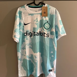 Inter Milan bortatröja Nike M - Inter Milan bortatröja från Nike i storlek M. Tröjan har ett unikt kartmönster i ljusblått och vitt, med klubbmärke och Nike-logga på bröstet. Digitalbits och Lenovo som sponsorer. Tillverkad i lätt och ventilerande polyester, perfekt för match eller träning.