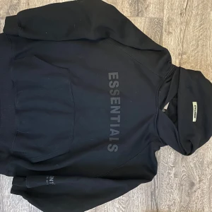 Svart Essentials hoodie med tryck - Svart hoodie från Essentials med stor logga tryckt framtill. Klassisk känguruficka, huva och ribbade muddar. Perfekt för en chill och avslappnad stil. Gjord i mjuk bomullsblandning för extra komfort.