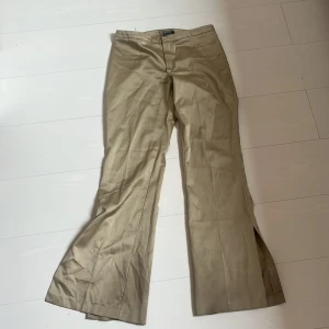 Beiga vida byxor från Cam i bomull - Snygga beiga/ guldiga byxor från Cam med låg midja, vida ben och slits nertill. Byxorna har en klassisk look med hög midja och är tillverkade i mjuk bomull. Perfekta för dig som gillar en relaxed och trendig stil.