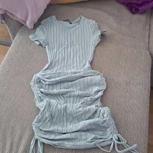 Ljusgrå ribbad klänning med snörning - Superfin ljusgrå klänning med ribbat tyg och snörning i sidorna. Klänningen har korta ärmar och rund halsringning, samt snygga rynkade detaljer längs sidorna som ger en cool och trendig look.