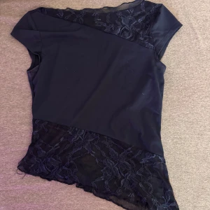 Mörkblå topp med spetsdetaljer - Snygg mörkblå topp med korta ärmar och transparenta spetsinslag upptill och nertill. Asymmetrisk nederkant och blommig spets ger en cool och elegant vibe. Perfekt för dig som gillar unika detaljer och stilren look.