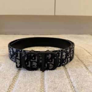 Svart och vit Dior bälte med monogram - Säljer ett snyggt Dior bälte med svart och vit monogramdesign och stor CD-logga i matt svart metall. Bältet är brett och har en cool, modern look. Materialet är textil med skinn på insidan. Perfekt för att lyfta vilken outfit som helst.