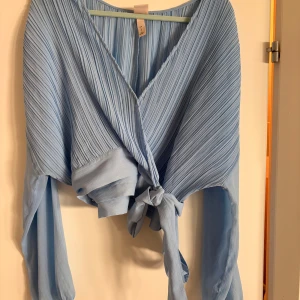 Ljusblå omlottopp från H&M - Superfin ljusblå omlottopp från H&M med plisserat tyg och långa ärmar. Toppen har v-ringning och knyts snyggt i midjan.
