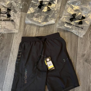 Svarta träningsshorts Under Armour M - Svarta träningsshorts från Under Armour med elastisk midja och snörning. Snyggt logotryck på benet och diskret mönster i regnbågsfärger längs sidan. Lätta och luftiga, perfekta för gymmet eller löprundan.