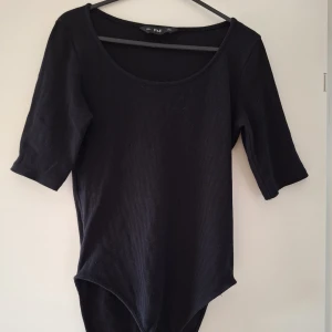 Svart ribbad body från F&F - Svart ribbad body från F&F med rund halsringning och halvlånga ärmar. Kroppsnära passform och tryckknappar i grenen för enkel på- och avtagning. Perfekt att styla med jeans eller kjol för en clean look.