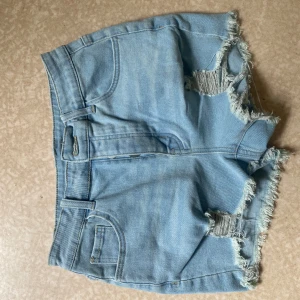 Ljusblå jeansshorts med slitningar - Snygga ljusblå jeansshorts med fransiga kanter och slitna detaljer framtill. Klassisk femficksmodell med bälteshällor och dragkedja. Perfekta för varma dagar när du vill ha en avslappnad och trendig look.