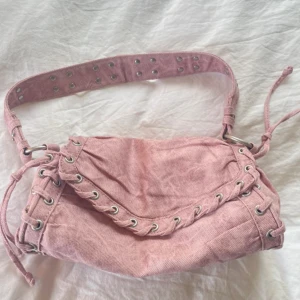 Rosa handväska från Zara -  Rosa axelväska i bomull med bred axelrem och snygga snörningsdetaljer längs kanterna. Väskan har silverfärgade metalldetaljer och en innerficka med dragkedja. Perfekt för dig som vill ha en unik och trendig accessoar till din outfit. Väskan saknar en knapp(sista bilden)