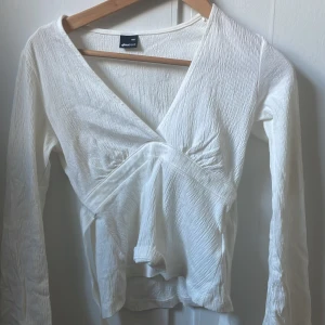 Vit omlottopp från Gina Tricot - Supersnygg vit omlottopp från Gina Tricot med lång ärm och v-ringning. Toppen har en lätt skrynklig struktur i tyget och sitter bekvämt med en lite lösare passform. Perfekt att styla med jeans eller kjol för en fräsch look.