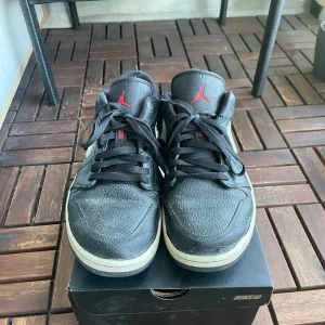 Nike Air Jordan 1 Low svart/röd, strl 46 - Säljer ett par Nike Air Jordan 1 Low i svart skinn med röda detaljer och vit sula. Skorna har klassisk Jordan-logga på plösen och en unik swoosh i vitt med penseldrag-look på sidan. Perfekta för dig som gillar streetstyle och sneakers med attityd.
