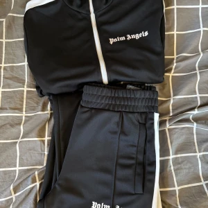 Palm Angels svart träningsset - Palm Angels svart träningsset med vit logga på både jacka och byxor. Setet har vita ränder längs ärmar och ben, dragkedja på jackan och elastisk midja på byxorna. Tillverkat i mjuk polyester för en sportig look. STORLEK M 