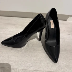 Svarta lackpumps från Nelly - Snygga svarta pumps i glansigt lackmaterial från Nelly. Skorna har spetsig tå och hög klack, vilket ger en elegant och trendig look. Perfekta för dig som vill sticka ut med klassiska men ändå moderna klackar.