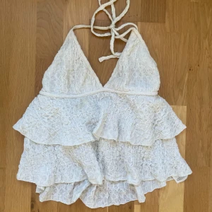 Vit volangtopp från Gina Tricot - Supersöt vit sommartopp med volanger och spetsliknande struktur. Toppen har knyt i nacken och v-ringad form, vilket ger en romantisk och feminin vibe. Perfekt för sommaren! Toppen har en liten fläck som syns på andra bilden!