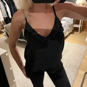 Svart linnetopp med spets från Samsa - Svart linnetopp från Samsa med tunna axelband och v-ringning dekorerad med spets. Snygg och enkel modell som är perfekt att styla med jeans eller kjol. 