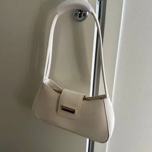 Beige axelväska i skinnlook - Snygg beige axelväska med stilren design och guldfärgad detalj på locket. Väskan har en mjukt rundad form och smal axelrem. Perfekt för dig som gillar minimalistisk stil och vill ha en väska som passar till det mesta.