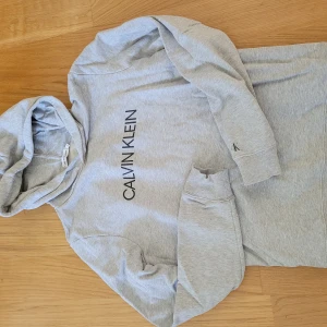 Grå hoodie från Calvin Klein Jeans - Snygg ljusgrå hoodie från Calvin Klein Jeans med svart logga framtill och liten 'ck'-brodyr på ärmen. Klassisk huva och ribbade muddar. Perfekt för en chill och stilren look.
