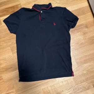 Pikétröja från U.S. Polo - Snygg svart pikétröja från Ralph Lauren med klassisk krage och röda detaljer på kragen och loggan. Tröjan har korta ärmar och knäppning framtill. Perfekt för en stilren och avslappnad look.  Pris går att diskutera!