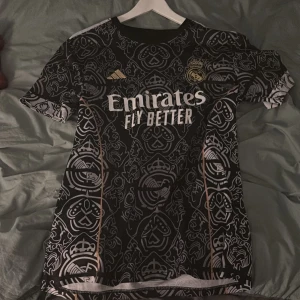 Real Madrid Mbappé tröja Adidas  - Säljer en svart Real Madrid fotbollströja från Adidas med Mbappé och nummer 9 på ryggen. Tröjan har korta ärmar, vit text och detaljer i guld samt ett unikt mönster över hela tyget. Materialet är lätt och andas, perfekt för match eller träning.