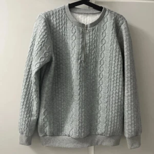 Ljusgrå kabelstickad tröja med half zip - Ljusgrå tröja med kabelstickat mönster och ribbade muddar. Tröjan har en snygg half zip-dragkedja med ringdetalj framtill. Tillverkad i mjuk polyesterblandning med lite stretch för extra komfort. Perfekt för kyliga dagar.