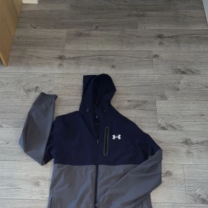 Blå och grå vindjacka från Under Armour - Snygg tvåfärgad vindjacka från Under Armour i marinblått och grått. Jackan har huva med dragsko, hel dragkedja framtill och en vertikal bröstficka med dragkedja. Perfekt för blåsiga dagar och sportiga outfits. Nypris 1,250, säljs inte längre 