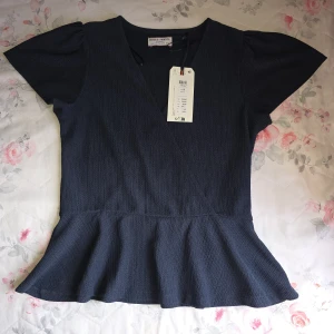 Mörkblå omlottopp från Holly & Whyte - Supersnygg mörkblå omlottopp från Holly & Whyte med korta puffärmar och volang nertill. Toppens struktur är lätt ribbad och den har en v-ringad hals. Perfekt för dig som gillar en feminin och trendig look. Helt ny med prislapp och oanvänd.