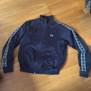 Svart track jacket från Fred Perry - Snygg svart track jacket från Fred Perry med dragkedja framtill och vita detaljer längs ärmarna. Jackan har ribbade muddar, ståkrage och två fickor. Perfekt för en sportig och stilren look.