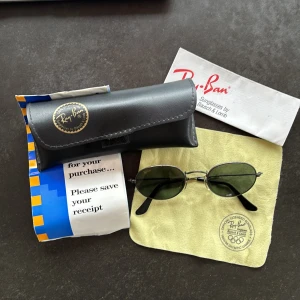 Ray-Ban ovala solglasögon i silver - Snygga vintage Ray-Ban solglasögon med ovala, mörkgröna glas och tunn metallbåge i silver. Klassisk och tidlös modell som ger en cool vibe. Kommer med originalfodral, putsduk och kvitto. Köpta 1995, sparsamt använda, allt i mycket gott skick. Perfekta för dig som vill ha en ikonisk look i solen.