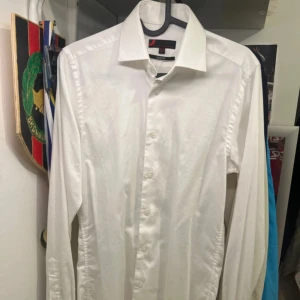 Skjorta  - Klassisk vit skjorta från Dressmann i slim fit-modell. Skjortan har lång ärm, knappar framtill och en stilren krage. Perfekt för dig som gillar en clean och enkel look. Storlek XS, passar dig som vill ha en smalare passform.