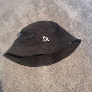 Svart bucket hat med LA-brodyr - Svart bucket hat i 100% bomull med vit LA-brodyr framtill. Klassisk form med bred brätte och små ventilationshål på sidorna. Perfekt för en chill streetwear-look och enkel att matcha med olika outfits.