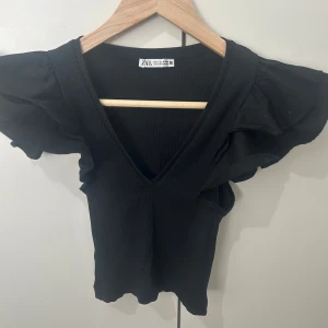 Svart ribbad topp med volangärm Zara - Supersnygg svart ribbad topp från Zara med djup v-ringning och stora volangärmar som ger en extra cool touch. Toppen är figurnära och har en stilren look som passar perfekt till jeans eller kjol. Materialet är mjukt och stretchigt för en bekväm känsla.