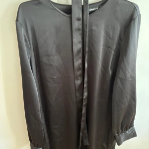 Svart satin-klänning från Gina Tricot - Stilren svart klänning i satin från Gina Tricot. Klänningen har långa ärmar med lätt puff vid ärmslut och ett smalt bälte i midjan för en snygg siluett. Perfekt för dig som gillar en enkel men elegant look.