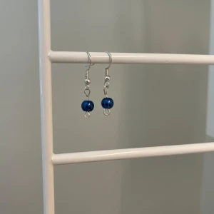 Silvriga örhängen med blå pärla - Ett par stilrena örhängen med silverfärgade krokar och en rund, blå pärla som hänger längst ner. Perfekta för att addera en färgklick till din look. Enkla men ändå unika och lätta att matcha med olika outfits.