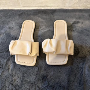 Beige sandaler med puffig rem - Trendiga sandaler i beige med fyrkantig tå och bred, puffig rem över foten. Tillverkade i syntetmaterial med platt sula, perfekta för varma dagar. Minimalistisk och modern design som ger en cool vibe till din outfit.