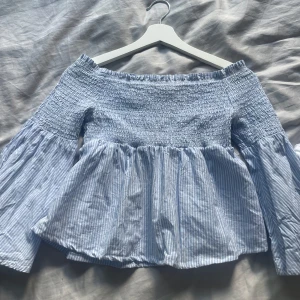 Off-shoulder blus - Supersöt offshoulder blus från Gina Tricot i blått och vitt med smock upptill och vida, långa ärmar. Blusen har tunna vertikala ränder och en luftig, flowy passform. Perfekt för vår och sommar när du vill ha en chill men ändå snygg look.