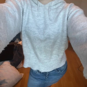 Ljusgrå stickad hoodie med huva - Supermjuk ljusgrå stickad hoodie med huva och dragsnören. Tröjan har långa ärmar och ribbade muddar vid ärmslut och nederkant. Perfekt för mysiga dagar och chill outfits.