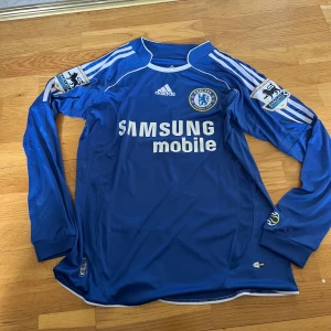 Chelsea långärmad Drogba 11 tröja - Chelsea FC långärmad fotbollströja med Drogba 11 på ryggen. Blå med vita detaljer, Adidas-logga och klubbmärke på bröstet. Premier League-patch på ärmarna och Samsung Mobile som sponsor. Tillverkad i lätt och ventilerande material.