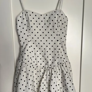 Vit prickig kortklänning med smala axelband - Supersöt vit kortklänning med svarta prickar och smala axelband. Slutsåld och eftertraktad! Ny med prislapp 
