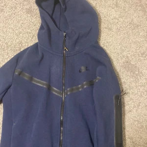 Mörkblå Nike tech fleece Nike - Säljer en mörkblå hoodie från Nike med dragkedja och huva. Jackan har ett diskret Nike-logotyptryck på bröstet och en svart randdetalj över bröstet. Perfekt för en sportig look.finns bara en skada där med Zipen har ändrat färg som ni ser på bilden det guldiga men man kan ta av.Finns några små små hål men man märker ej