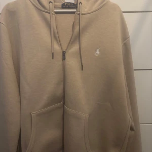 Beige hoodie från Polo Ralph Lauren - Snygg beige hoodie från Polo Ralph Lauren med dragkedja framtill och klassisk huva med snörning. Tröjan har två stora fickor och den ikoniska broderade loggan i vitt på bröstet. Perfekt för en chill och stilren look.  Den är helt ny 