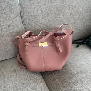 Rosa axelväska med gulddetaljer - Snygg rosa axelväska från H&M i mjukt, strukturerat syntetmaterial. Väskan har justerbar axelrem, guldfärgat spänne och öppet huvudfack. Perfekt storlek för det viktigaste och en stilren look med modern touch.