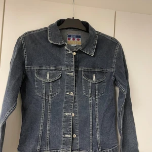 Mörkblå jeansjacka från BEST - Klassisk mörkblå jeansjacka från BEST med kontrastsömmar, två bröstfickor med lock och knappar framtill. Jackan har en rak passform och långa ärmar, perfekt för dig som vill ha en tidlös denimlook.