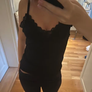 Svart linnetopp med spetskant - Säljer en svart linnetopp med smala axelband och snygg spetskant längs urringningen. Toppen är figurnära och har en enkel, stilren look som passar perfekt till jeans eller kjol. Materialet känns mjukt och följsamt mot huden.