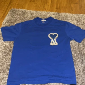 Blå t-shirt från AMI Paris med hjärtdetalj - Snygg blå t-shirt från AMI Paris med ikonisk hjärtformad A-logga i vitt på bröstet. Klassisk passform och rund halsringning. Tillverkad i mjuk bomull, perfekt för en clean och stilren look.