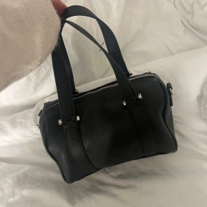 Svart handväska från Bizz - Snygg svart handväska från rizzo i klassisk boxig modell. Väskan är gjord i strukturerat skinn med dubbla handtag och silverfärgade metalldetaljer. Stängs med dragkedja upptill och har stilrena sömmar som ger en clean look. Använd fåtal gånger 