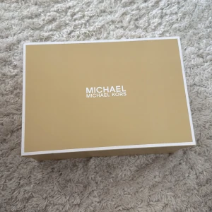 Michael Kors skor - Snygga sneakers från Michael Kors i beige och vitt med MK-mönster på sidorna. Skorna har guldfärgad dragkedja framtill med logodetalj och är tillverkade i skinn och syntet. Perfekt för dig som vill ha en stilren och trendig look.