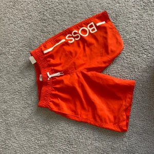 Röda badshorts från Hugo Boss XS - Snygga röda badshorts från Hugo Boss i storlek XS. Shortsen har vit BOSS-logga på ena benet, elastisk midja med vit snörning och innerbyxa i mesh. Perfekta för stranden eller poolen, riktigt fräsch färg och klassisk design.