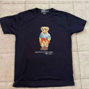 Svart Polo Bear t-shirt från Ralph Lauren - Svart t-shirt från Ralph Lauren med ikoniskt Polo Bear-tryck på bröstet. Klassisk passform med rund hals och korta ärmar. Trycket visar en björn i jeansjacka och röda detaljer. Perfekt statement-plagg för dig som gillar streetstyle.
