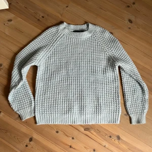 Ljusgrå stickad tröja från 157 - Supermysig ljusgrå stickad tröja från 157 med grov struktur och rund hals. Tröjan har långa ärmar och ribbade muddar vid ärmslut och nederkant. Lite nopprig men fortfarande i okej skick